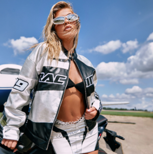 lady wearing sunglasses, leaning on a car wearing a black and white motocross jacket